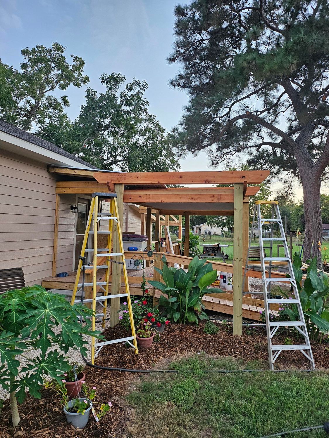 A wooden deck under construction next to a house with ladders and surrounding foliage.