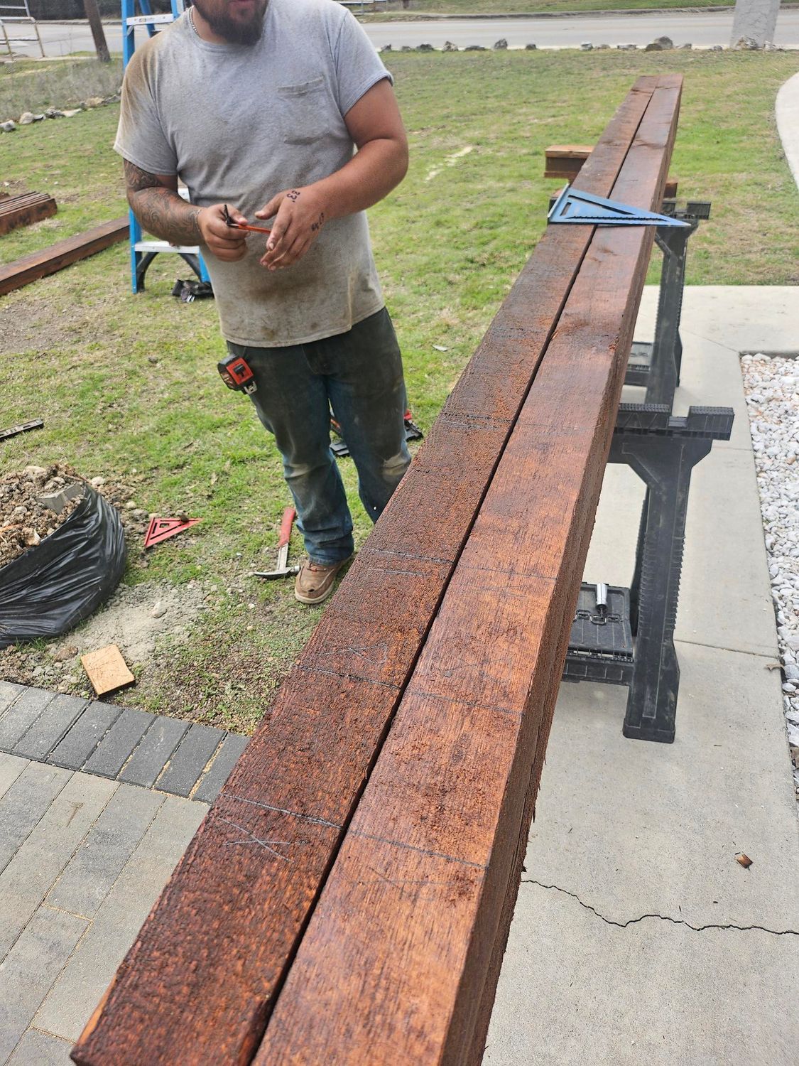 Man working on long, stained wooden beams on sawhorses outdoors.
