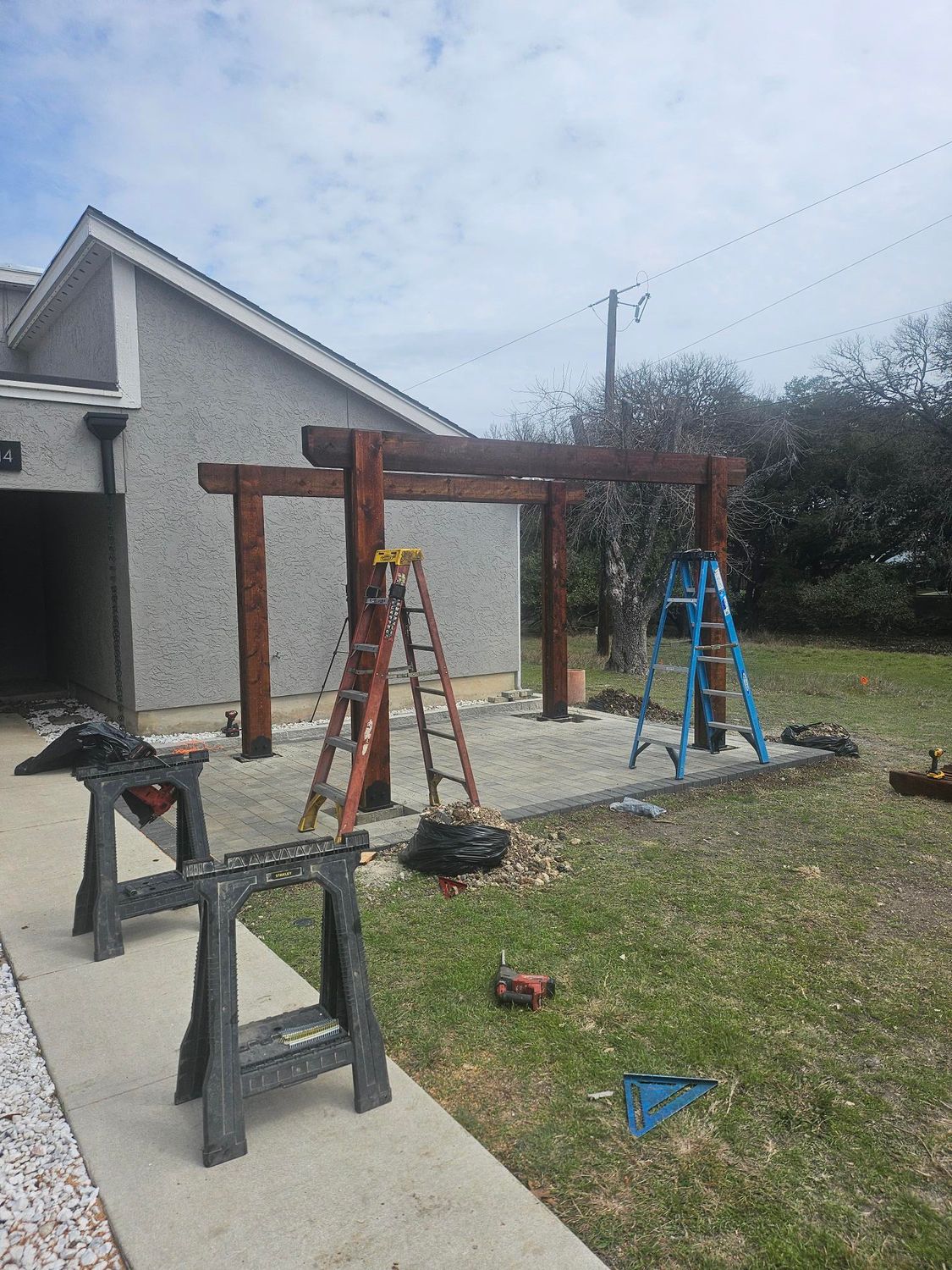 Construction of a wooden pergola on a concrete patio next to a house; ladders and tools present.