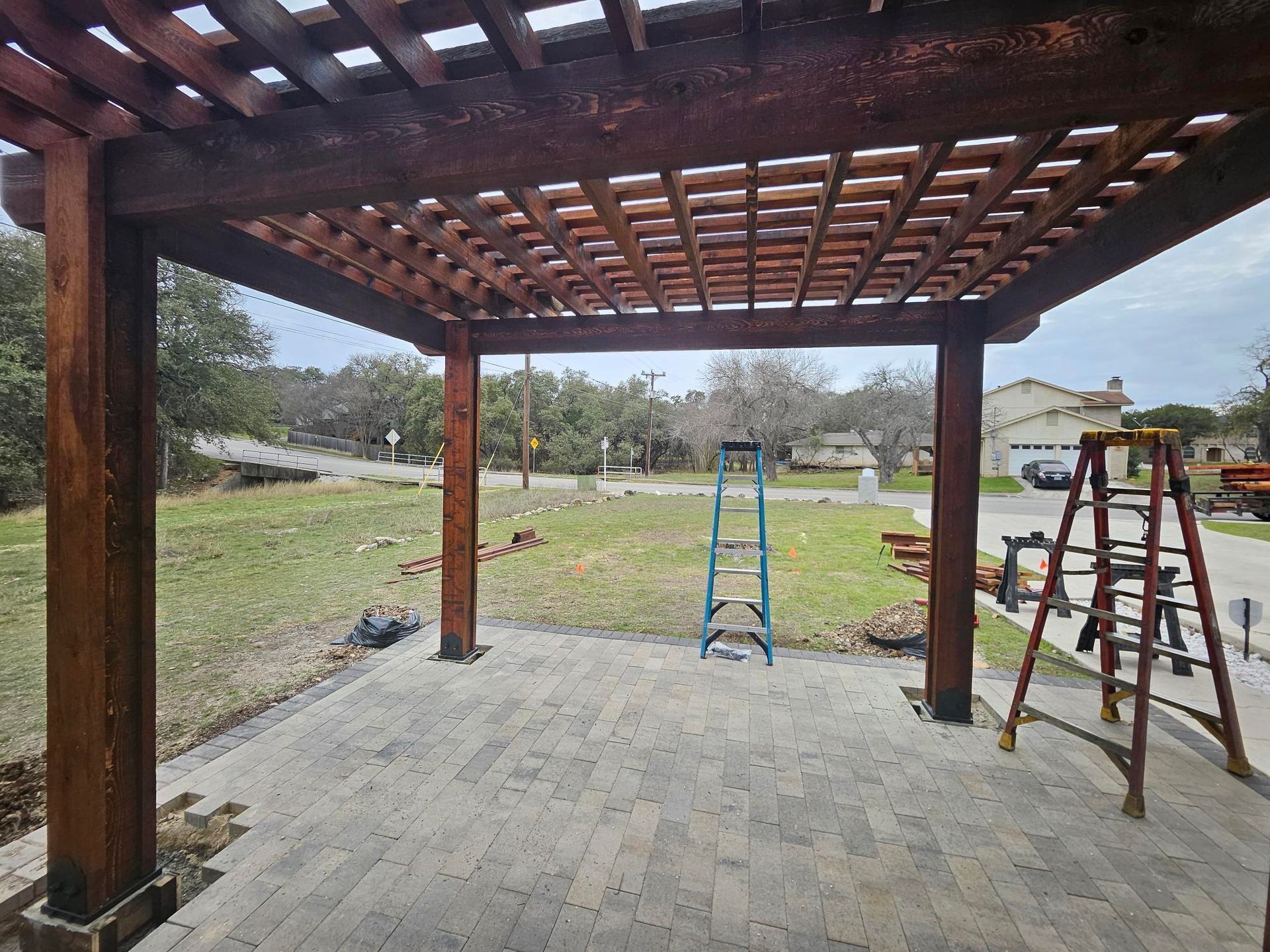 A pergola with a latticed roof over a brick patio, with a lawn and trees in the background. Ladders are visible.