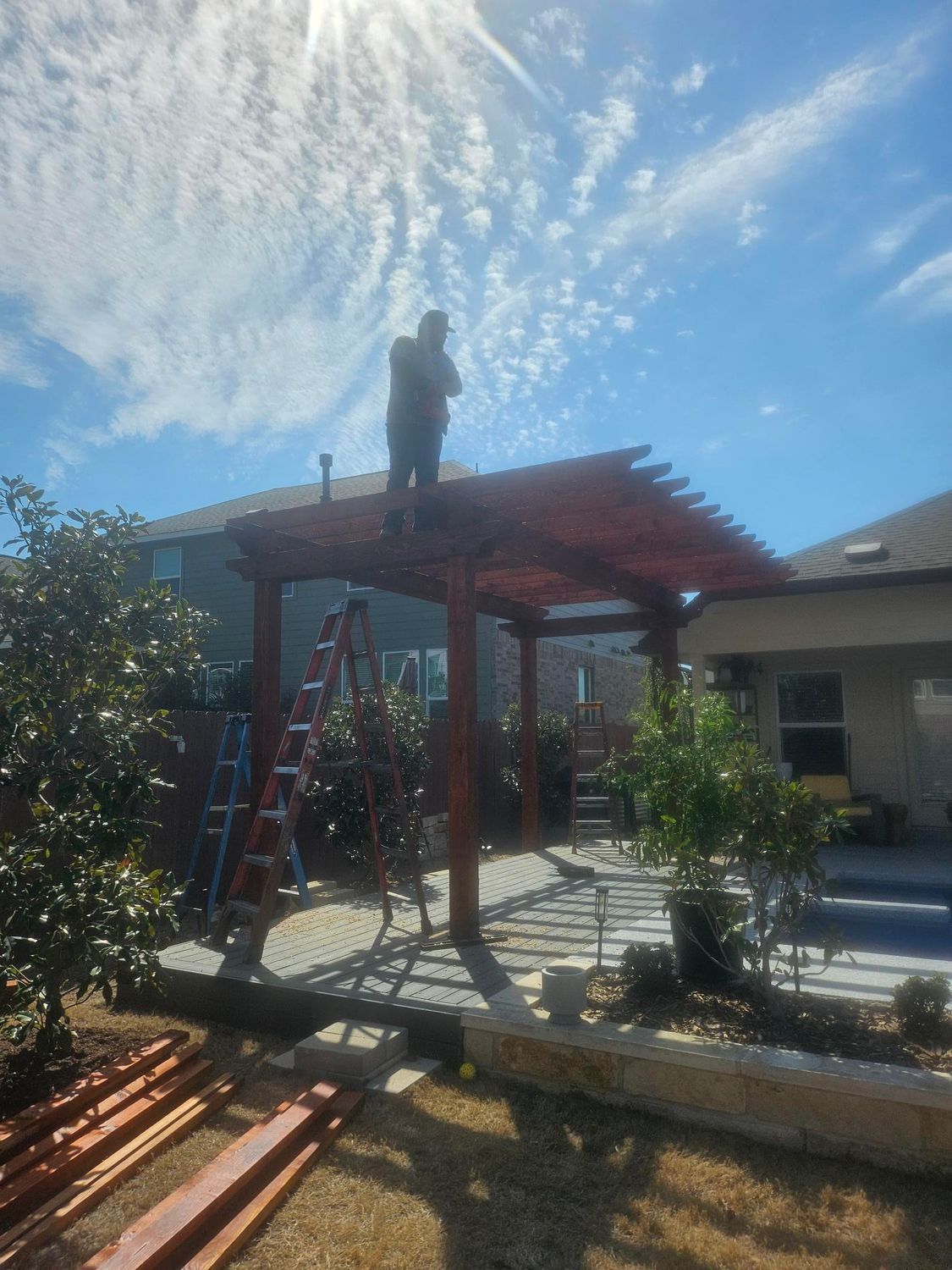 Person on top of a red pergola under a sunny sky; a ladder leans against it.