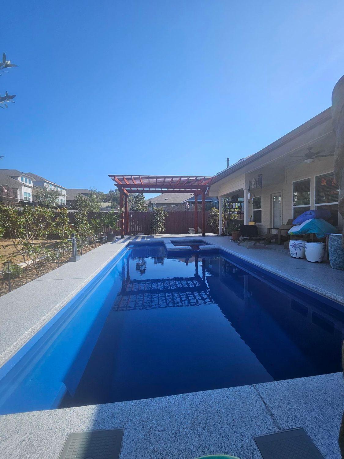 A rectangular swimming pool with dark blue water and a red pergola, next to a house under a clear, blue sky.