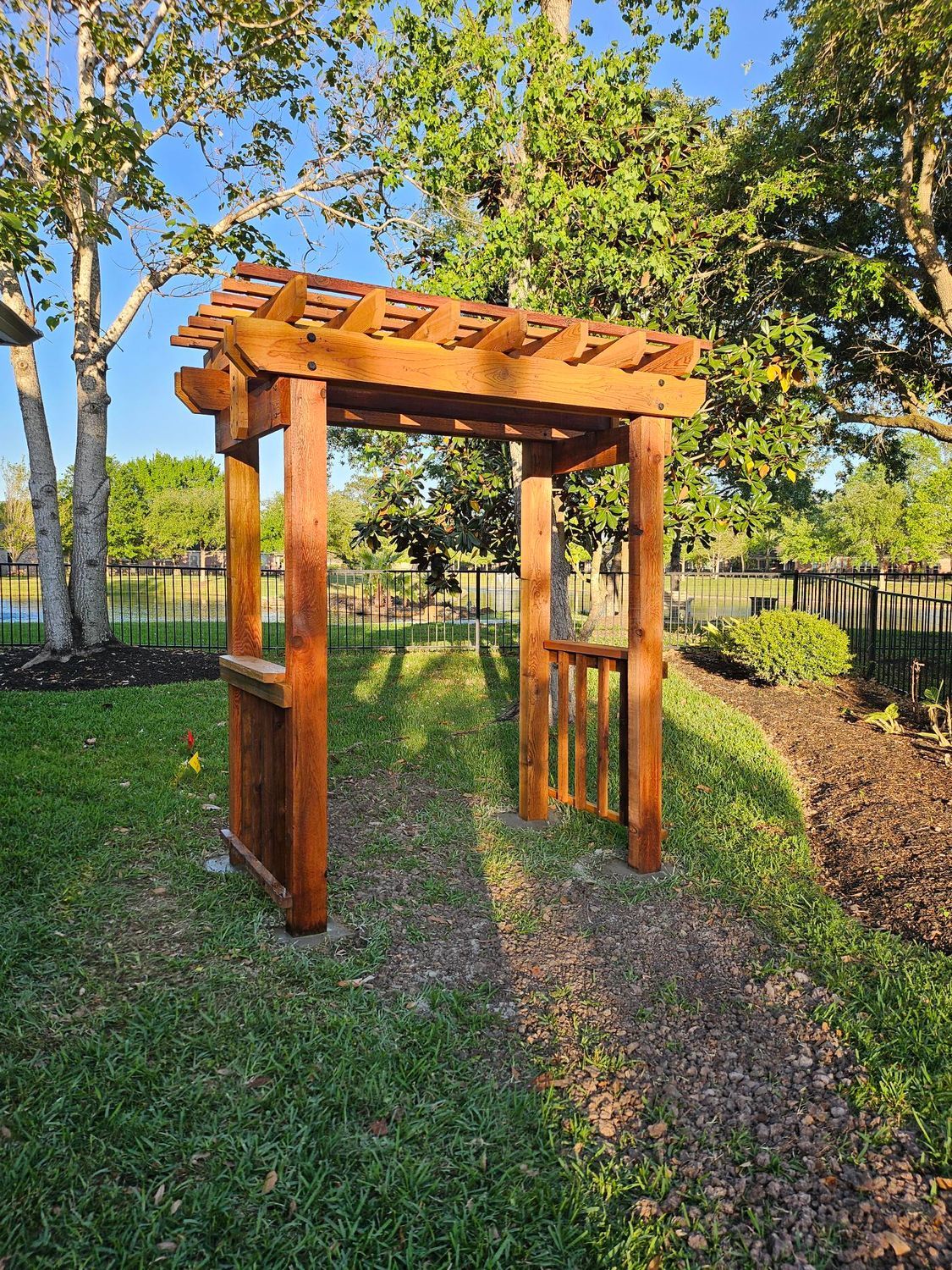 Wooden arbor with open gate on a grassy path, sunlight casts shadows.