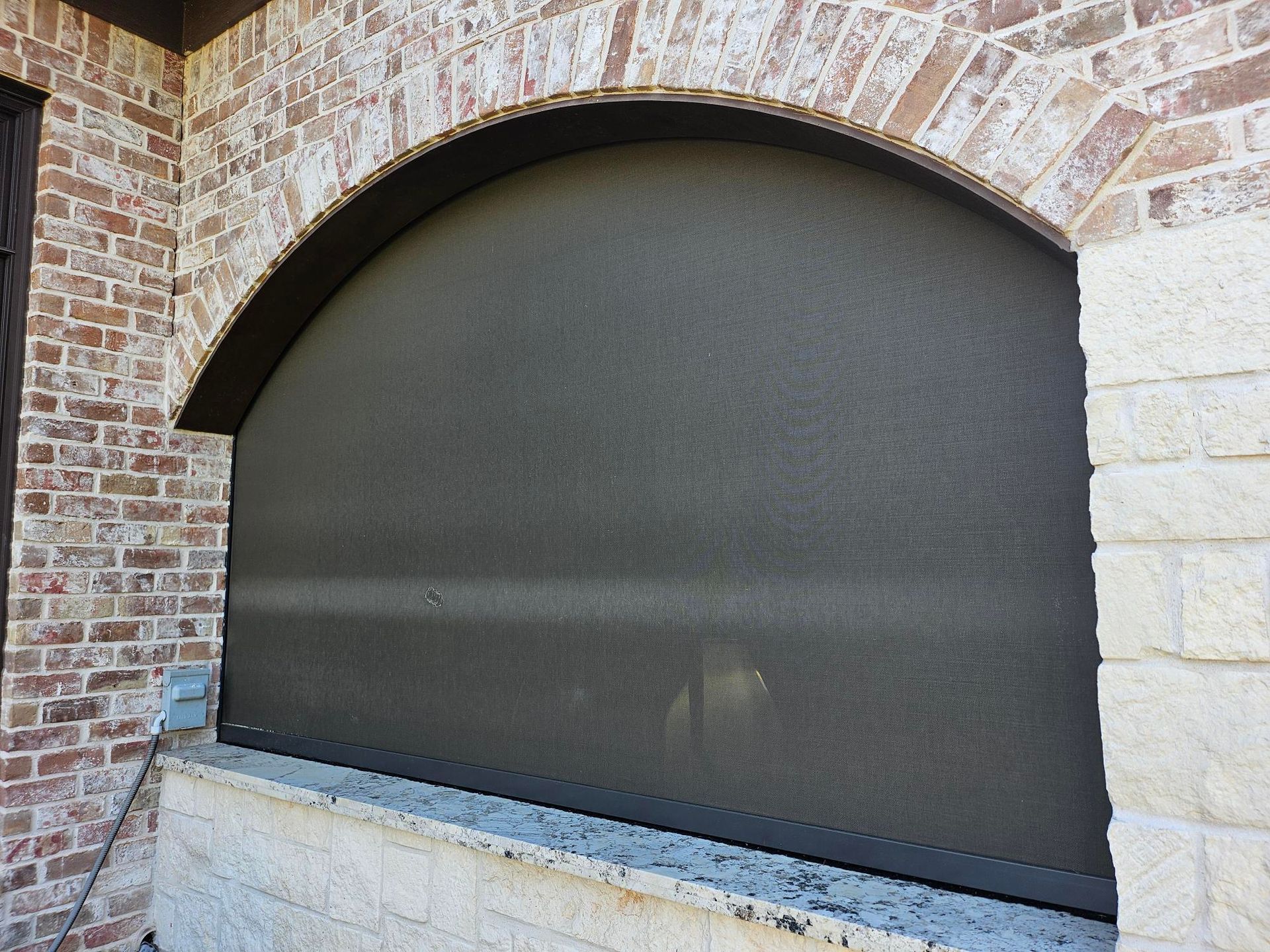 Exterior arched window covered by a dark, textured screen, set in brick and stone facade.