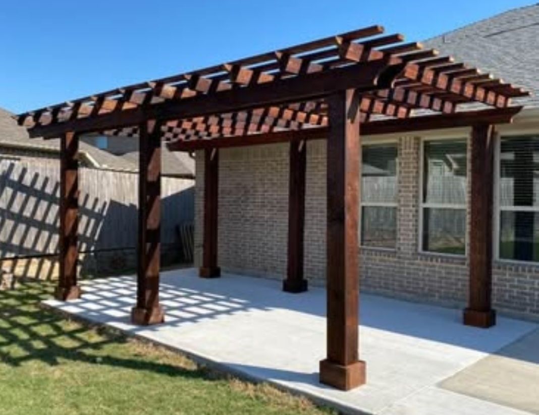 Brown wooden pergola over a concrete patio next to a brick house. Sunny day.