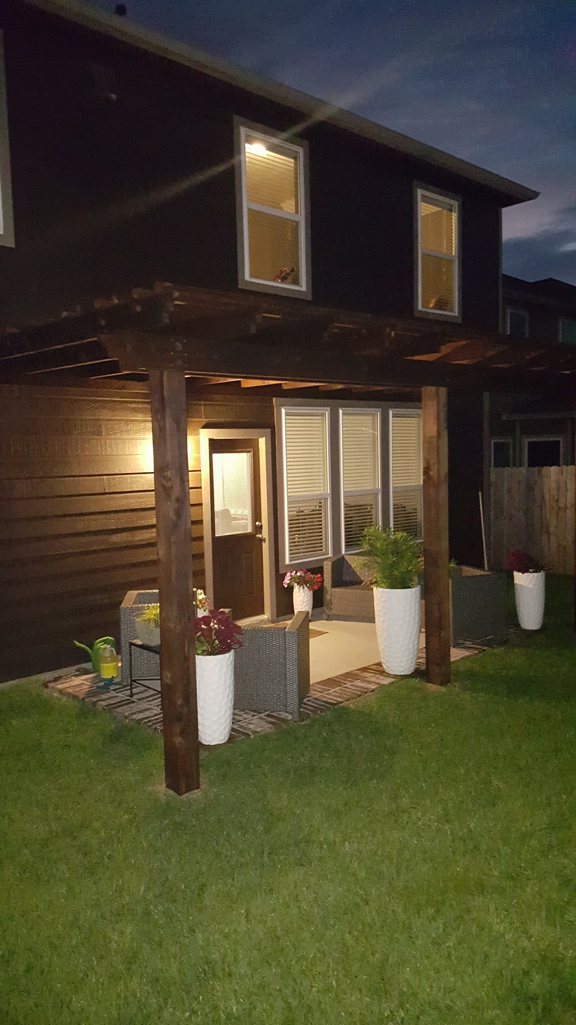 Backyard patio with pergola, dark siding, windows, door, potted plants, and green grass.