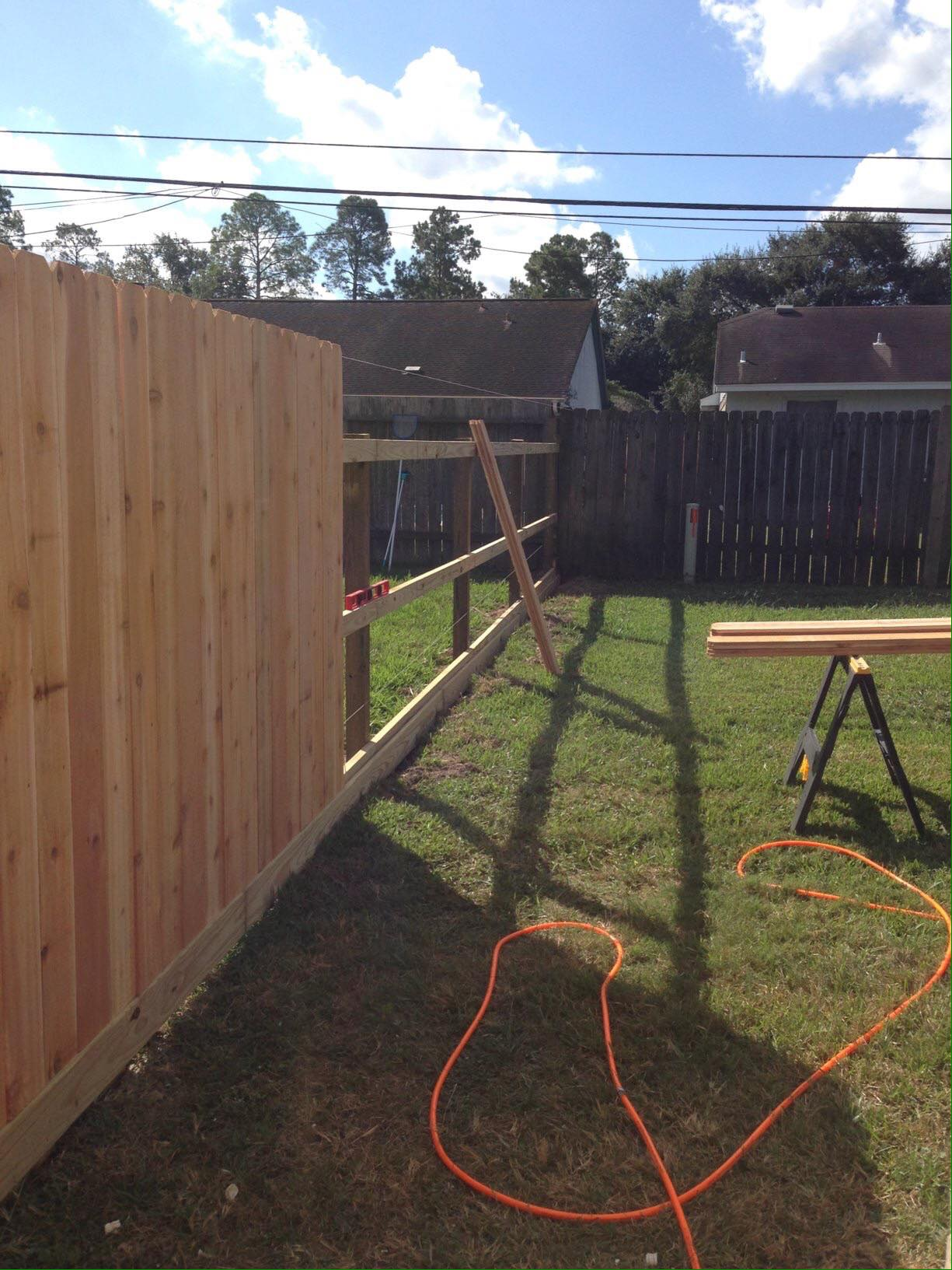 Wooden fence partially built in a grassy backyard, with tools and materials nearby on a sunny day.