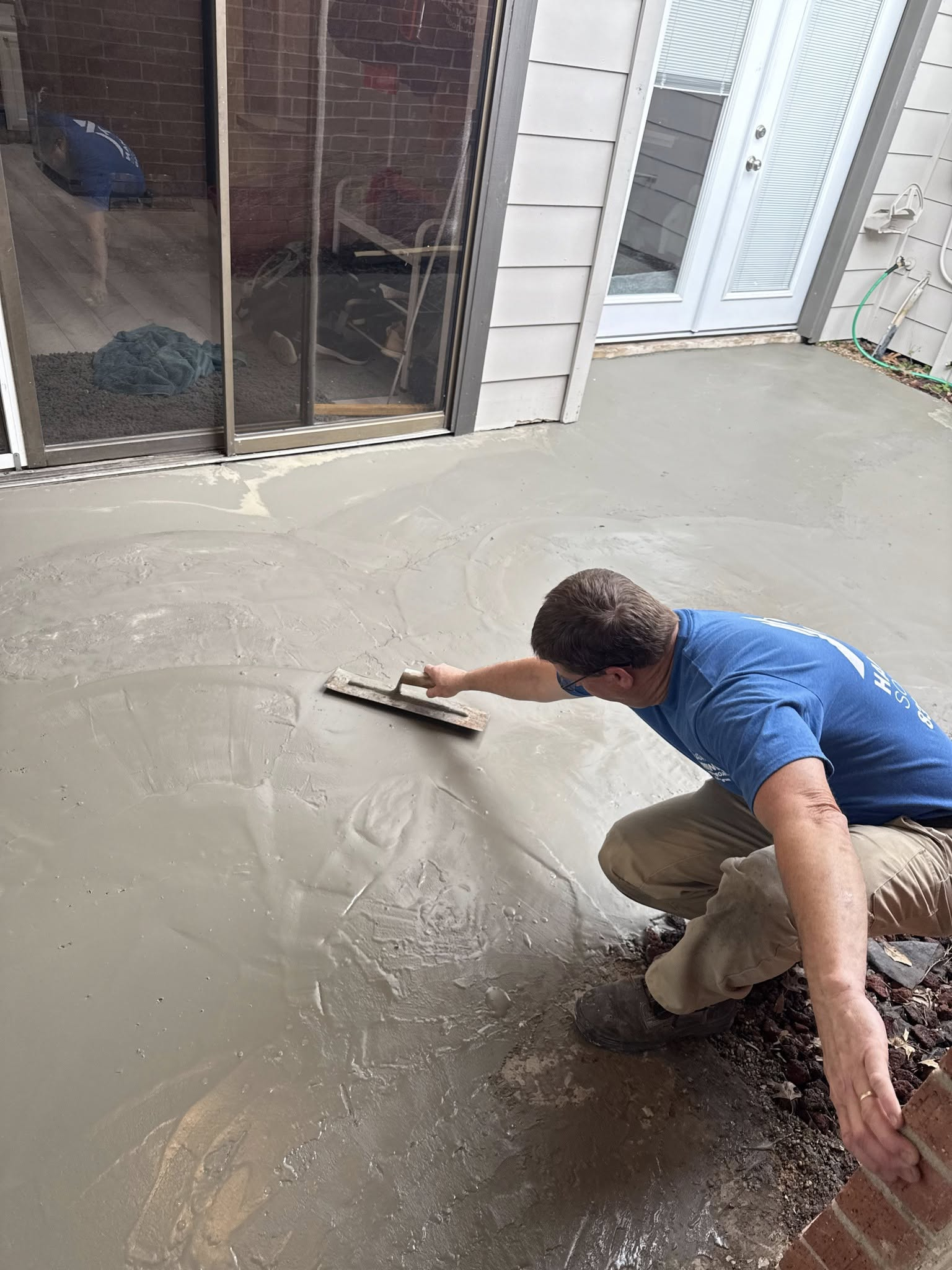 Person smoothing wet concrete with a trowel on a patio. Brick wall and sliding glass door are visible.