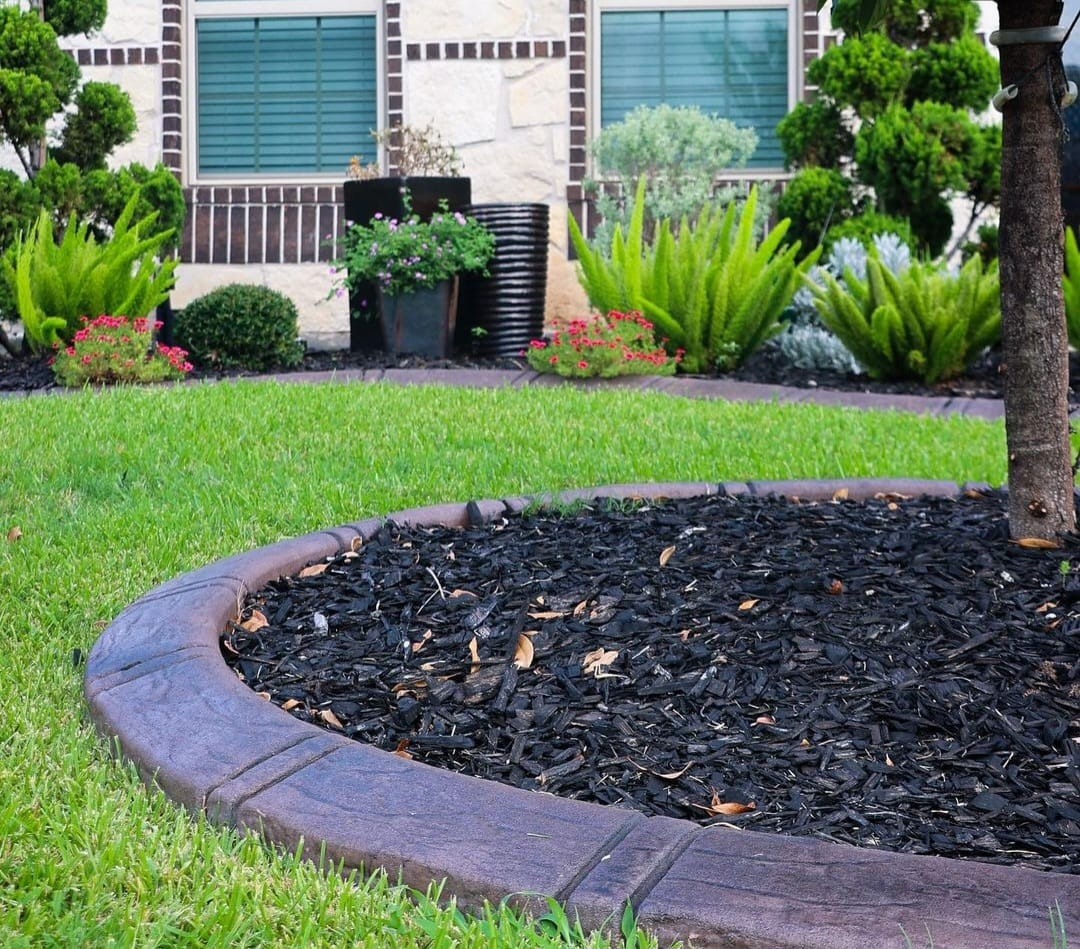 A landscaped front yard with green grass, a curved dark border, and dark mulch around plants near a house.