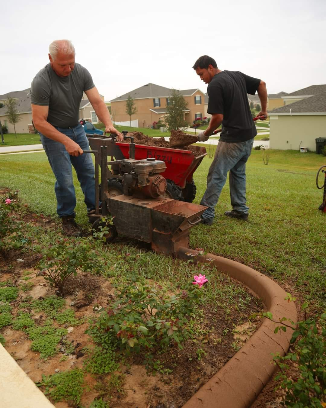 Two men installing drainage in a yard. One operates a machine, the other shovels dirt into a wheelbarrow. Brown corrugated tubing visible.
