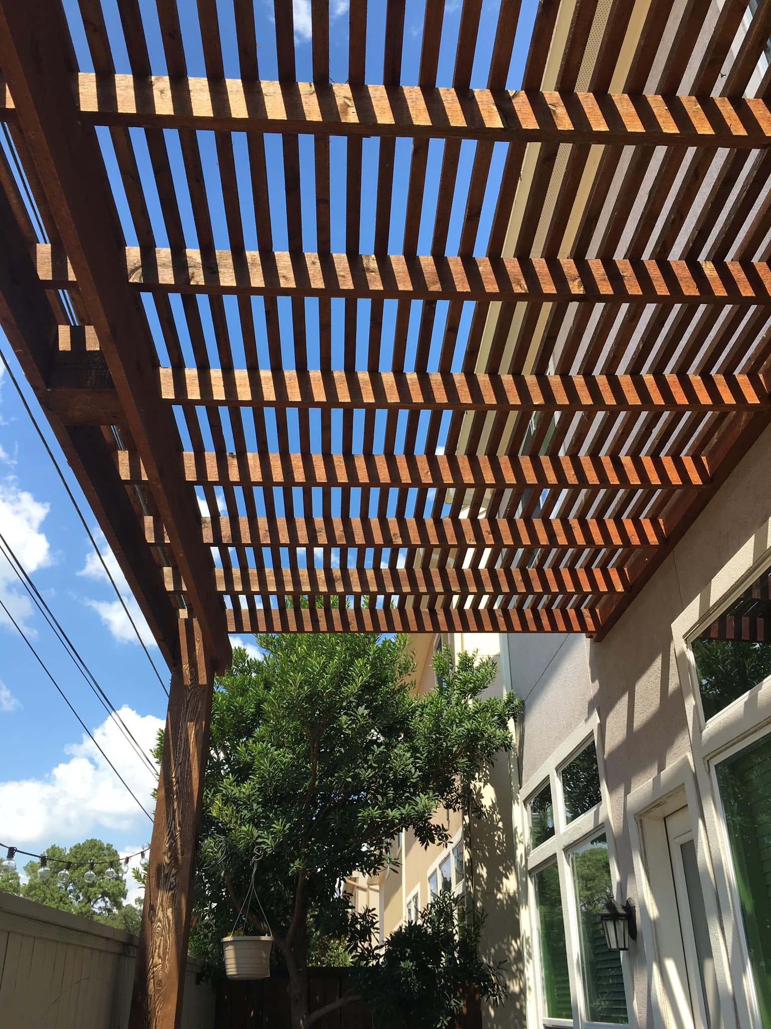 Wooden pergola over a patio, with blue sky visible through the slats; string lights on the left.