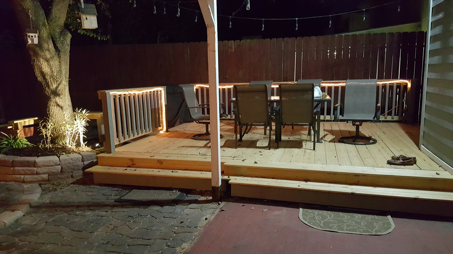 Wooden deck with illuminated railing and patio furniture at night. String lights and a tree are also visible.