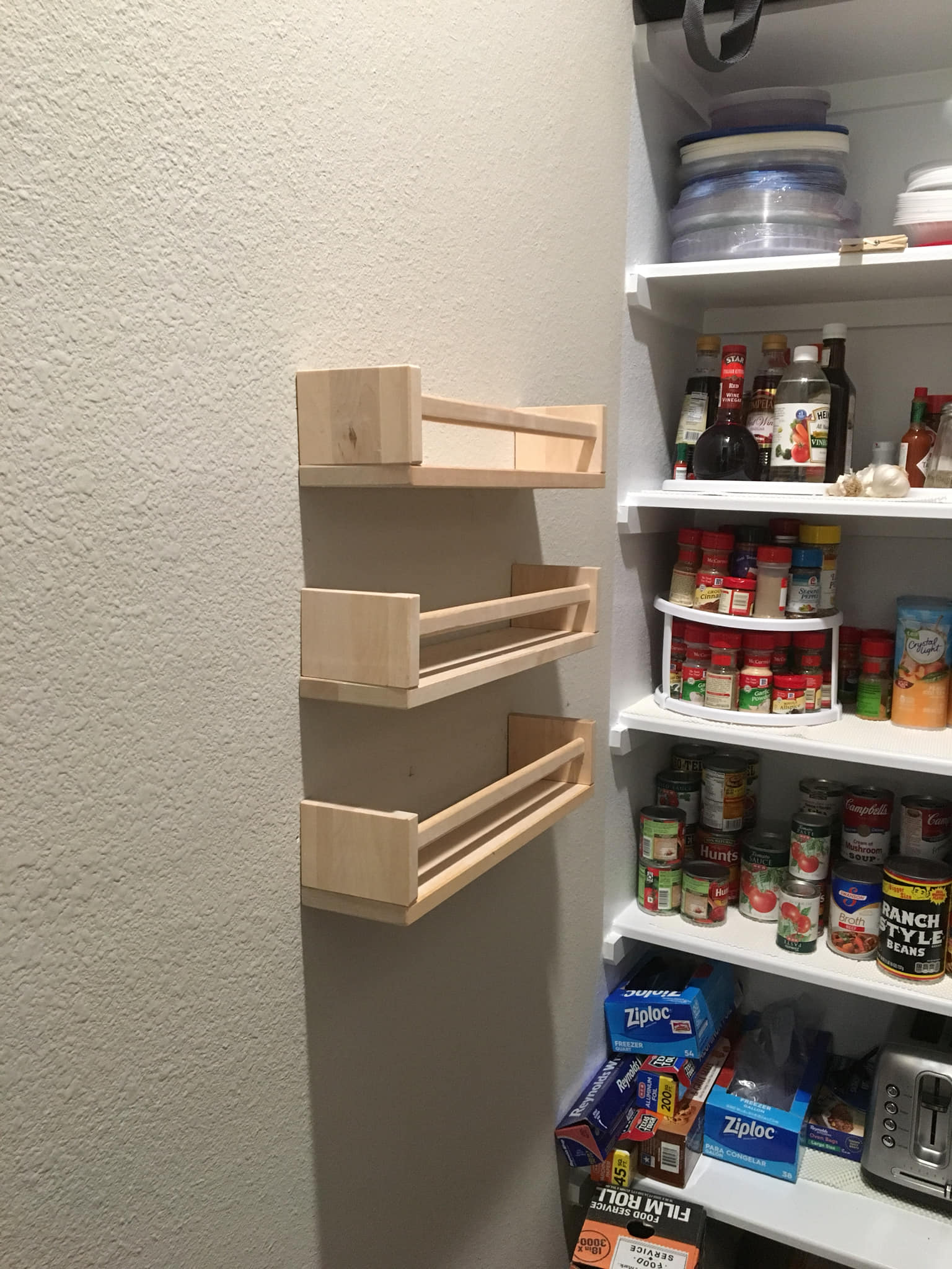Three light-wood spice racks on a textured wall, next to a pantry with shelves of food.