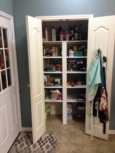 Open pantry with shelves filled with food items, adjacent to a white door. Aprons hang on the door.