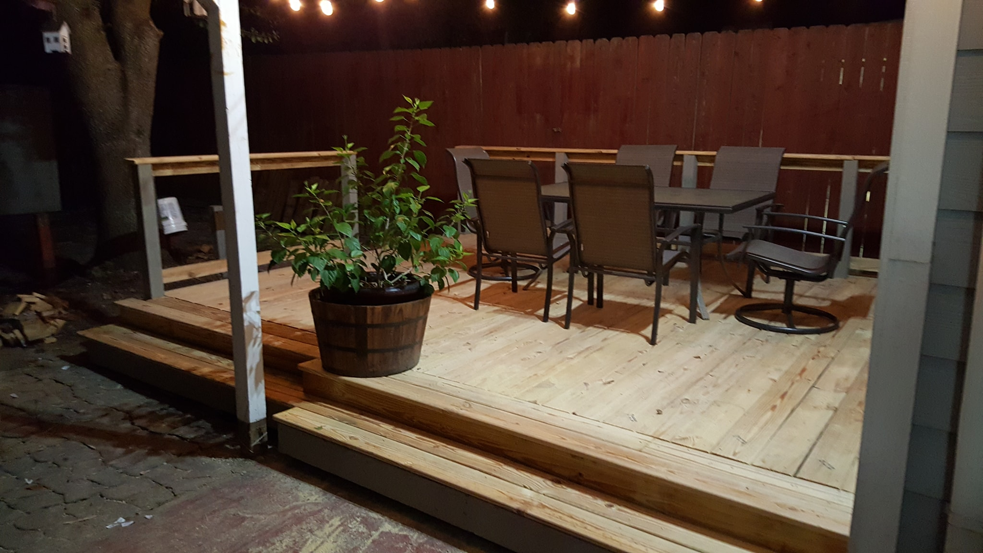 Wooden deck at night with a table, chairs, and potted plant, strung with lights against a dark fence.