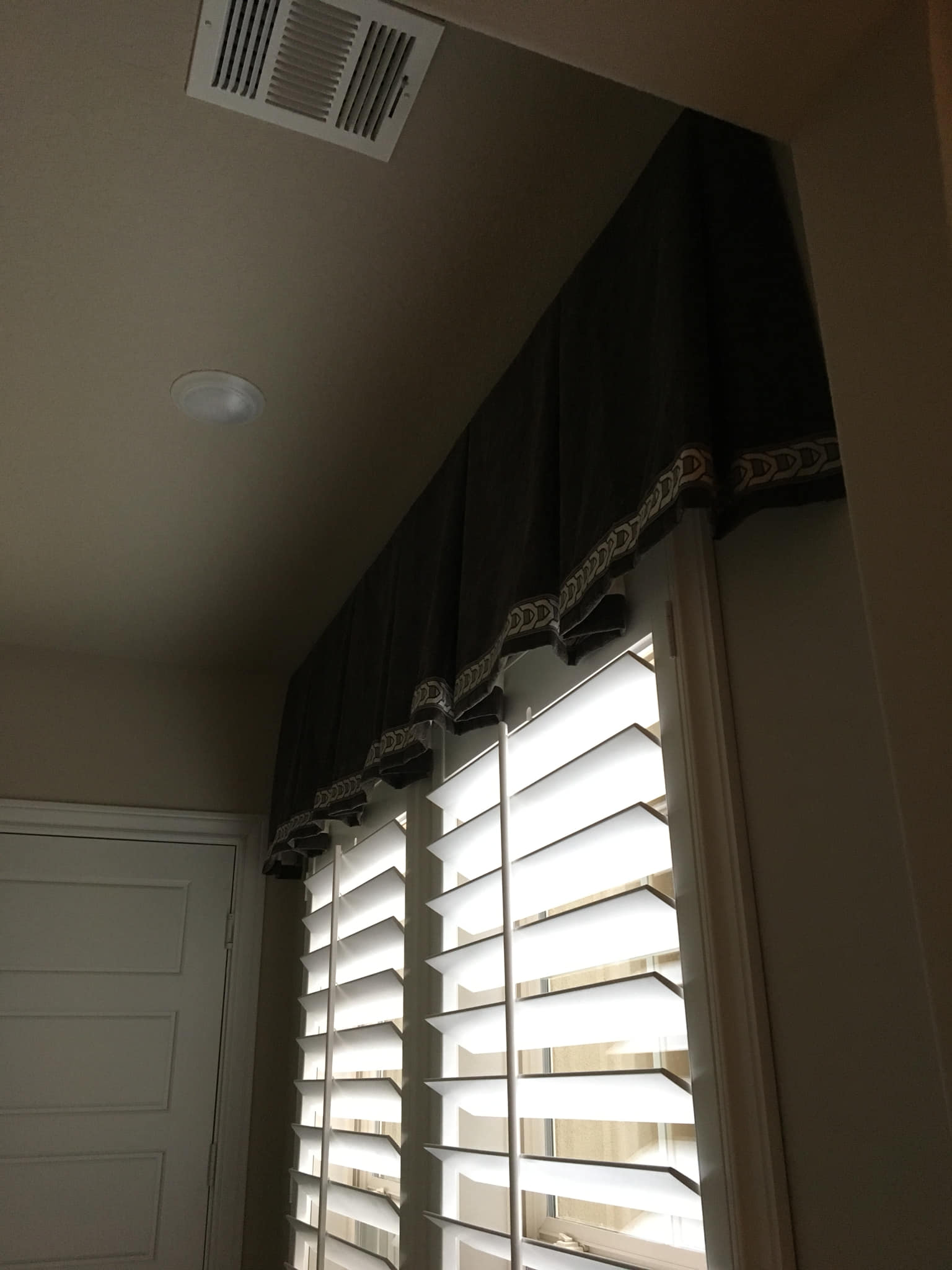 Dark valance with decorative trim above window shutters in a room.