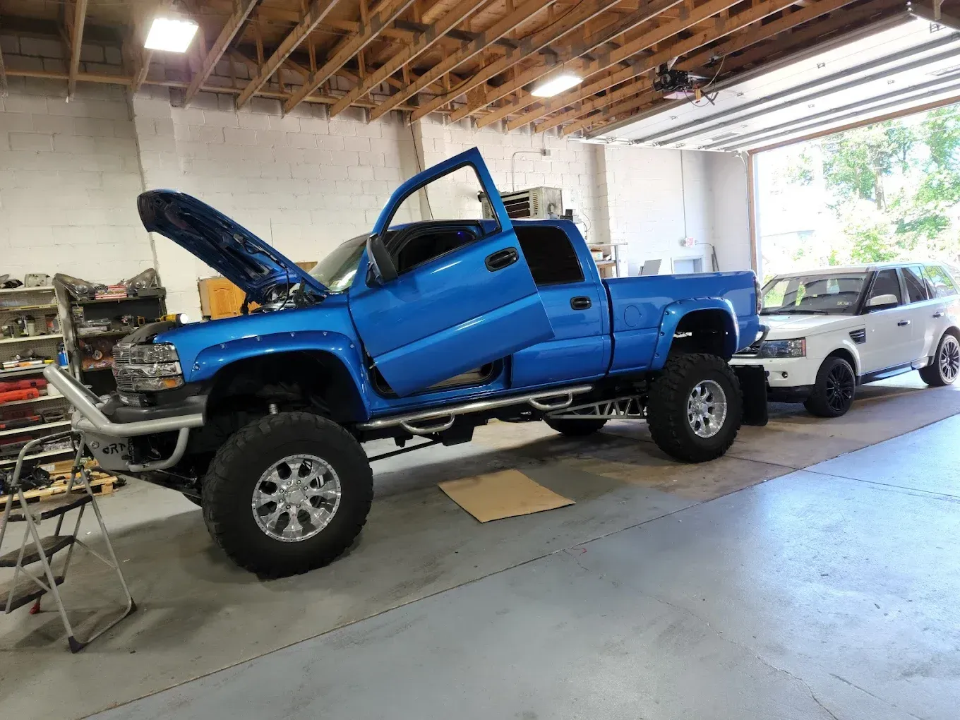 Blue lifted pickup truck with open hood and door inside a garage, next to a white SUV.