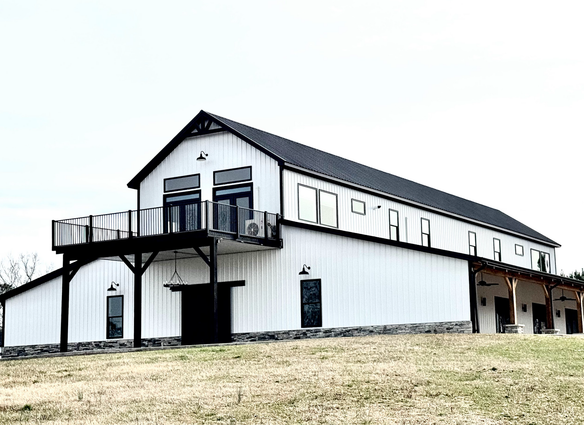 White barn with black roof, deck, and trim. Stone base. Set on a hill.
