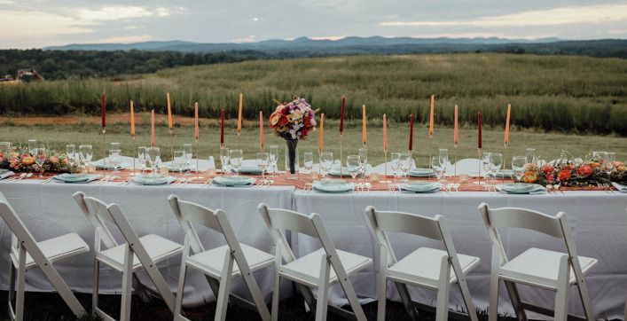 Long table set for a meal outdoors, white chairs, colorful floral centerpiece, candles, and a field in the background.