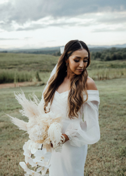 Bride in white dress holding a bouquet, looking down, standing in a field.