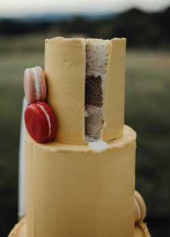 Yellow tiered cake with a slice removed, revealing layers. Red and pink macarons decorate.