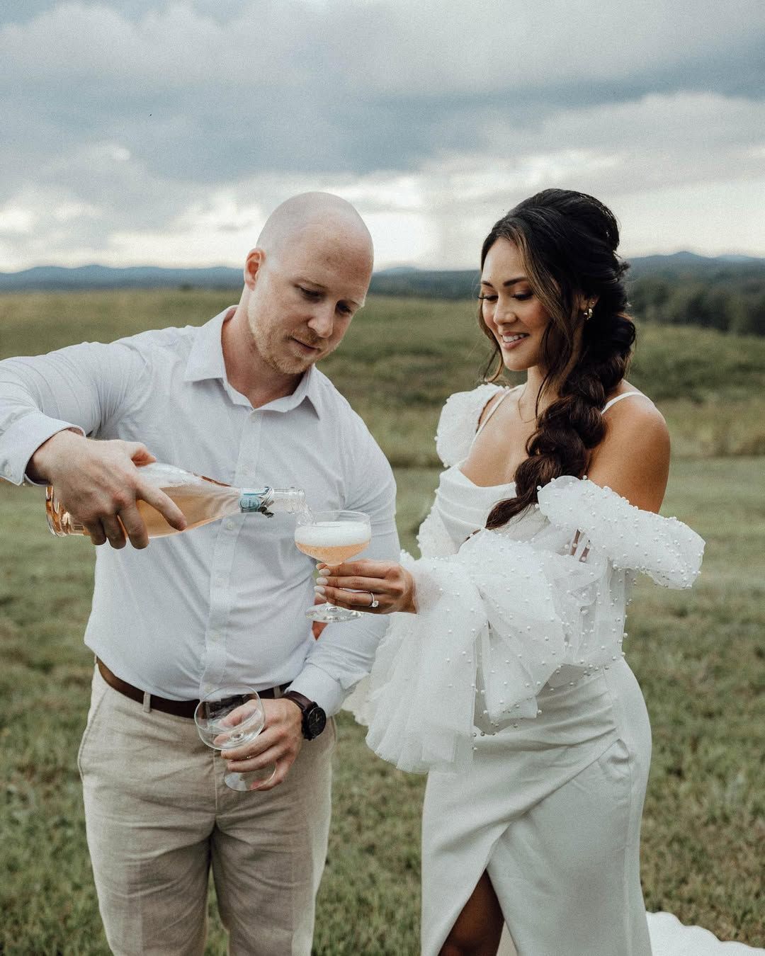 Man pouring champagne for a smiling woman in a wedding dress outdoors.