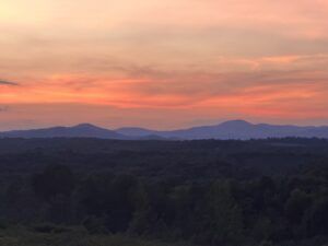 Sunset over tree-covered hills and distant mountains, with orange and pink sky.