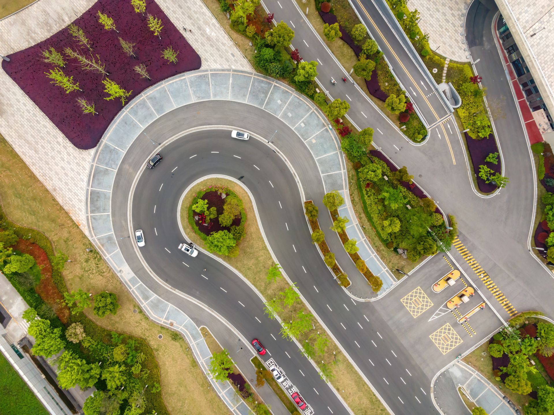 Aerial view of a complex road system with cars driving on looping curves, surrounded by green landscaping and trees.