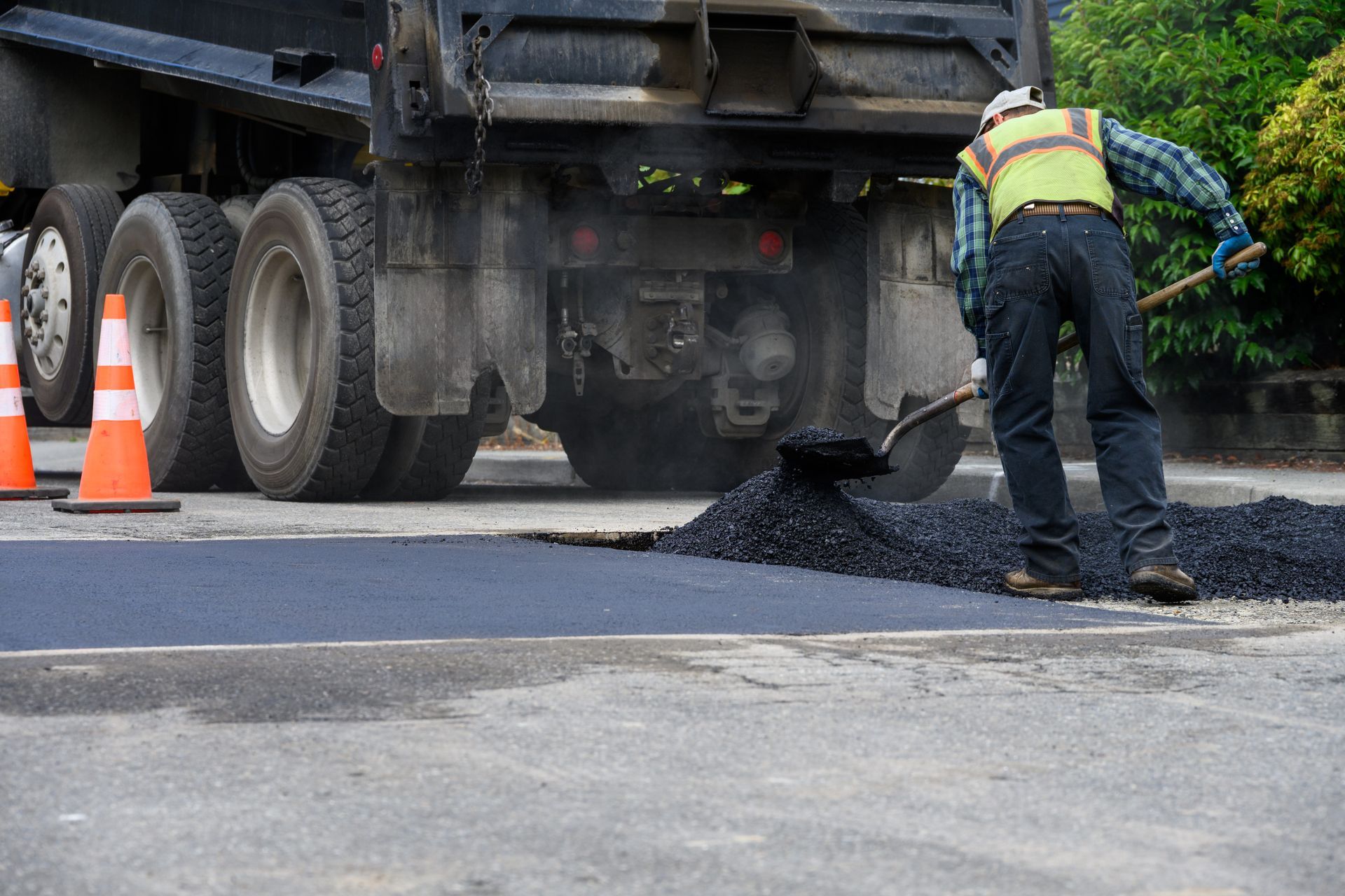 Worker spreading hot asphalt on a road surface near a dump truck and traffic cones.