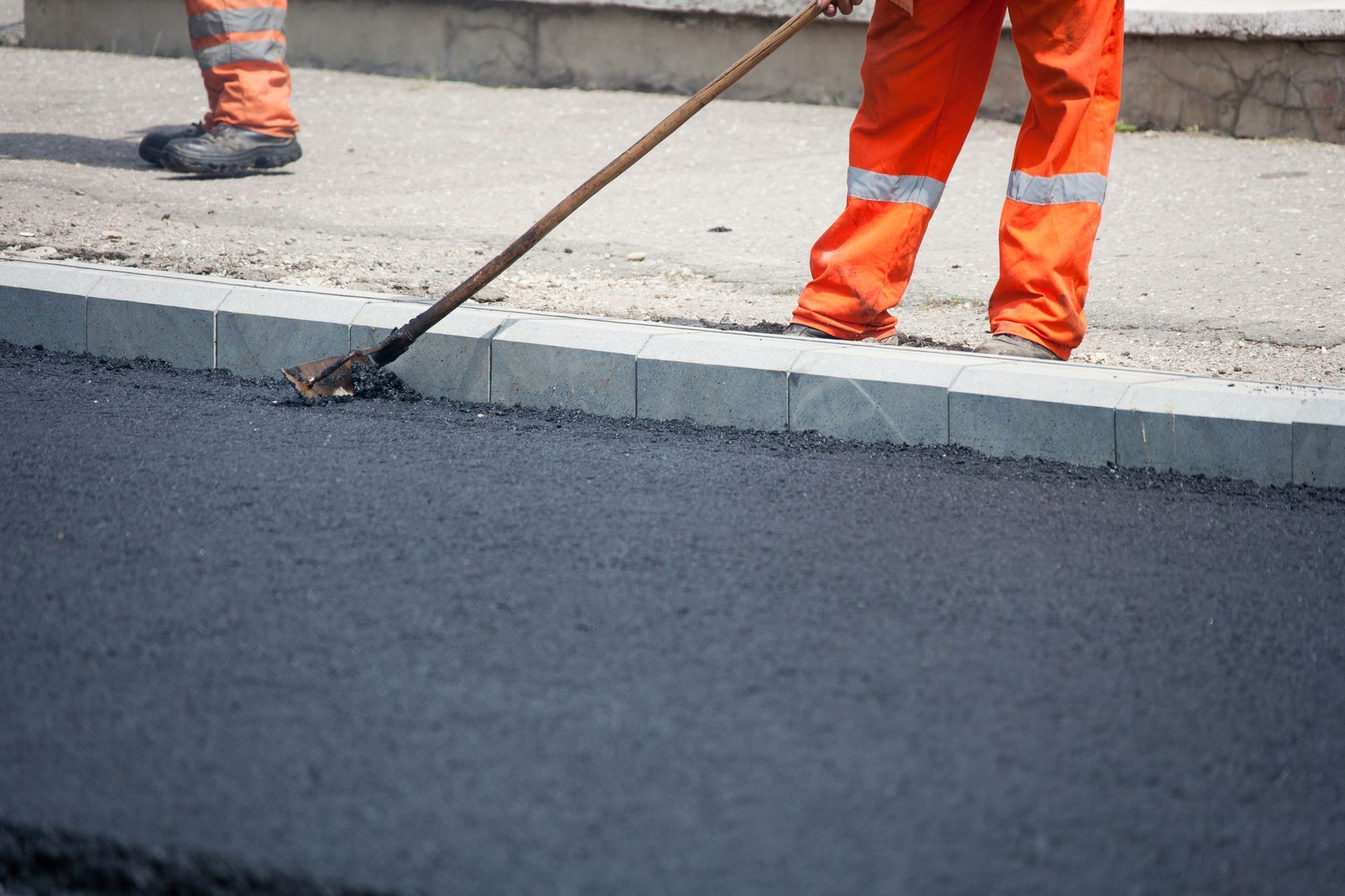 Road worker in orange overalls smoothing asphalt with a rake.