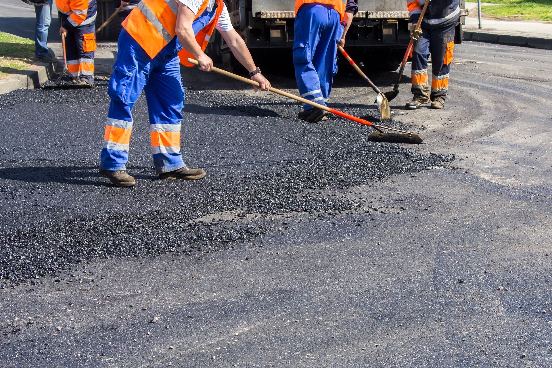Workers in high-visibility gear performing asphalt maintenance on a road with paving equipment.