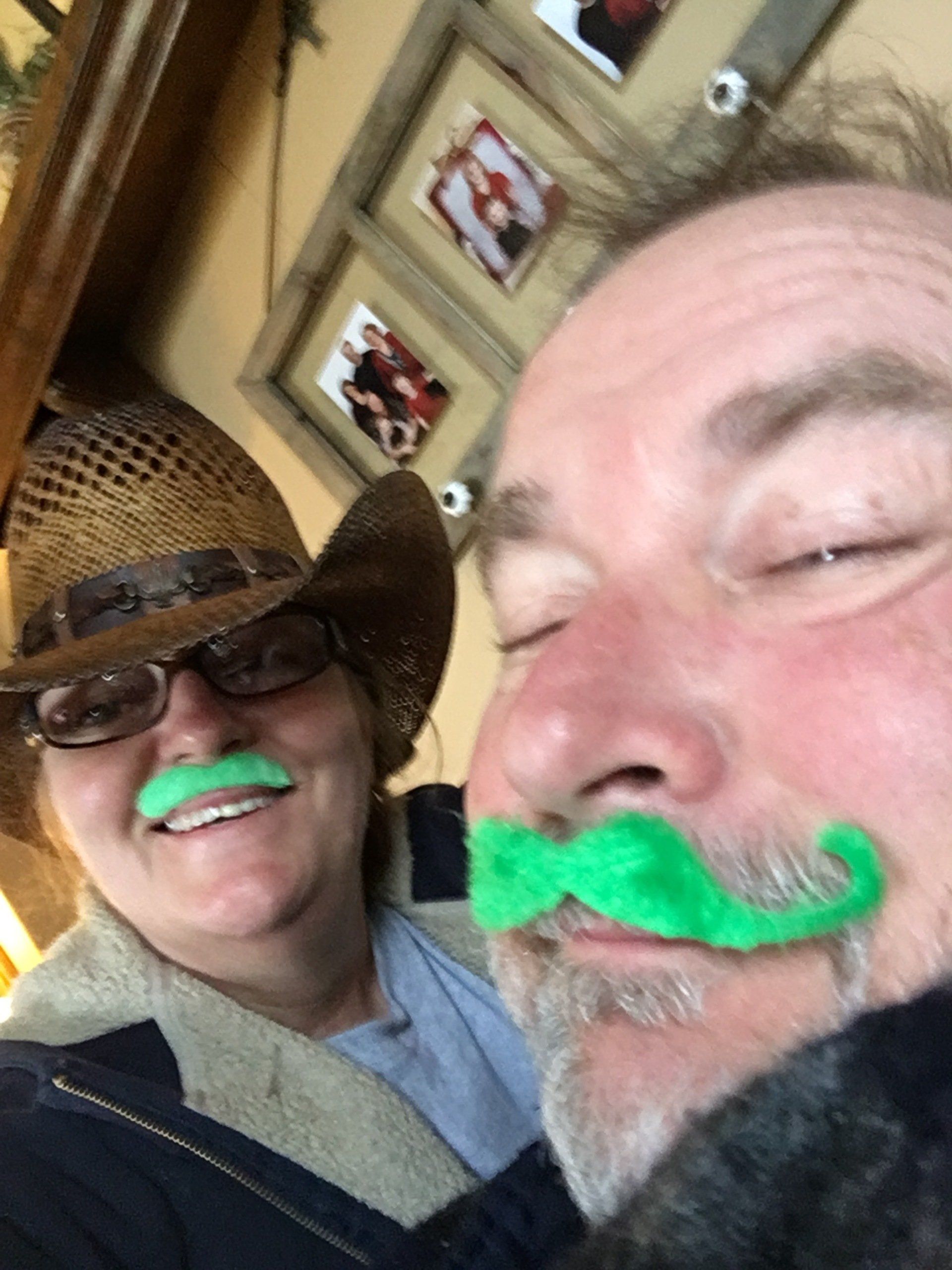 Two people wearing bright green faux mustaches pose for a selfie indoors with framed photos on the wall behind them.