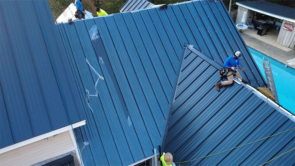 Roofer on a gray shingled roof, hammering shingles. Sunny day, house in background.