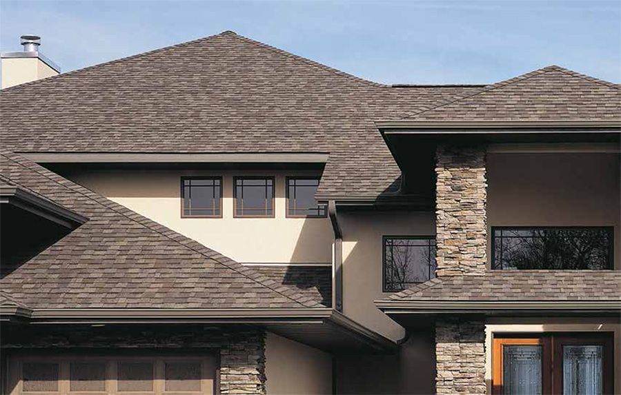 Roofer on a gray shingled roof, hammering shingles. Sunny day, house in background.
