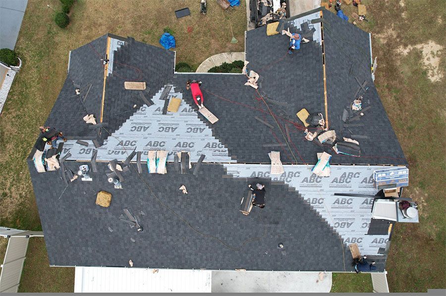 Roofer on a gray shingled roof, hammering shingles. Sunny day, house in background.