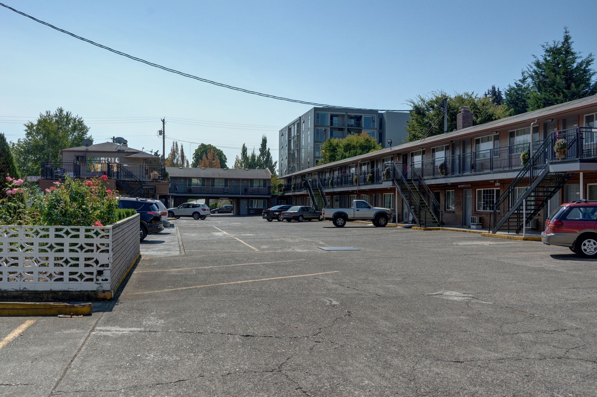A parking lot in front of a two-story motel with parked cars on a sunny day. A modern building is visible in the distance.