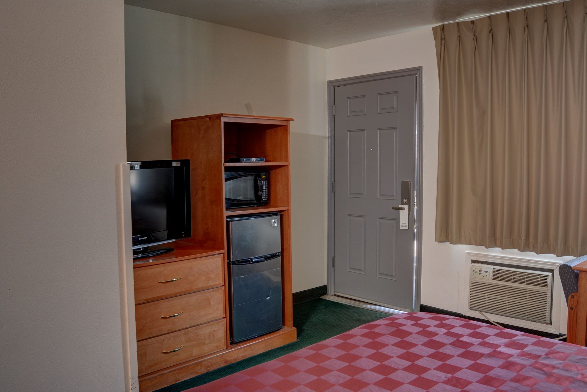 A hotel room interior with a bed, dresser with TV, microwave, and mini-fridge, and a closed door.