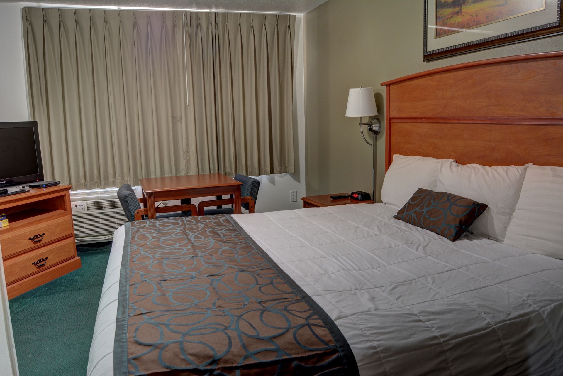 Hotel room interior with a bed, dresser, table, and window. A television sits atop the dresser.