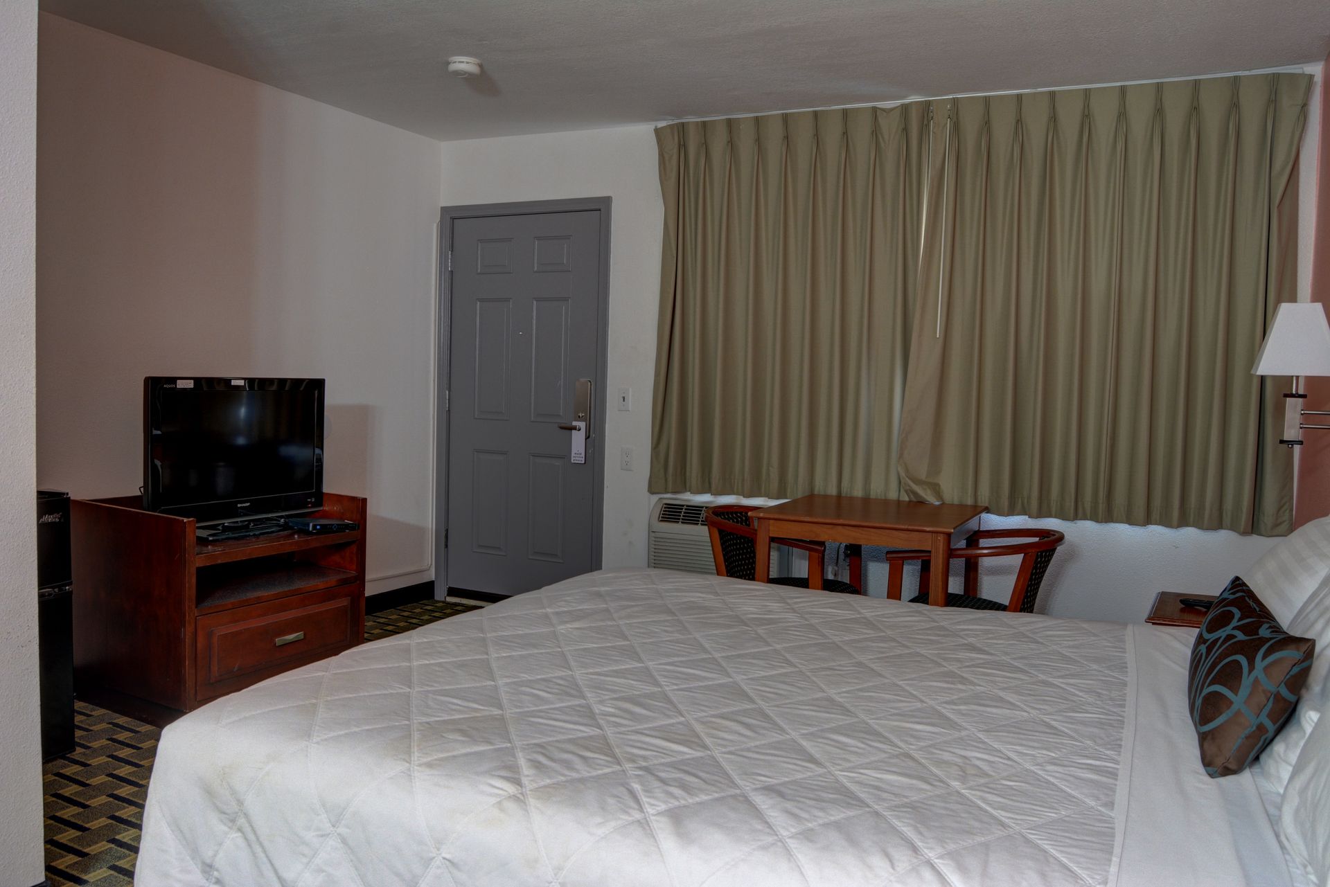 Hotel room with a bed, TV, small table, and closed door. Tan curtains cover a window.