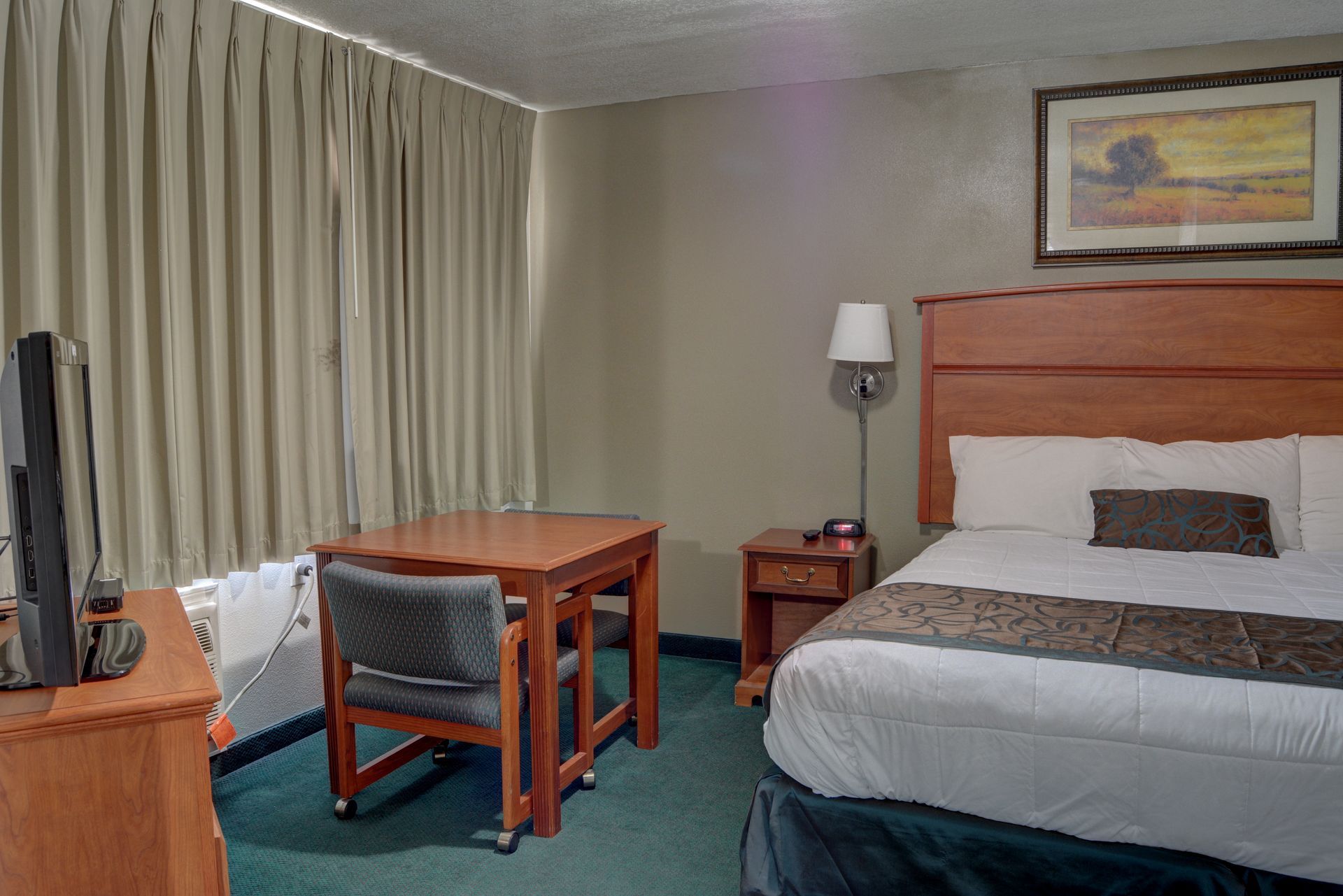 A hotel room with a bed, table with chair, TV, and window with curtains. The room has green carpet and earth-toned furniture.