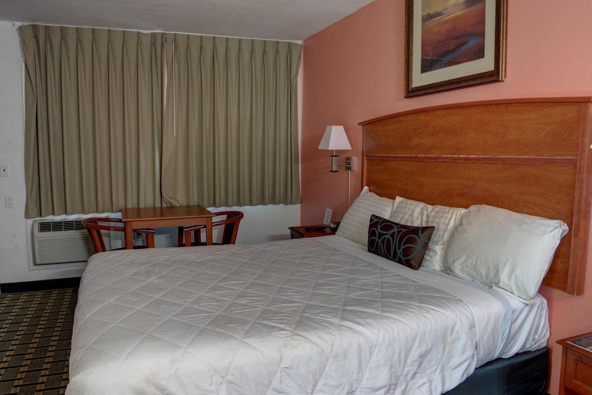 Hotel room with a queen-sized bed, wooden headboard, small table with two chairs, and a window with curtains.