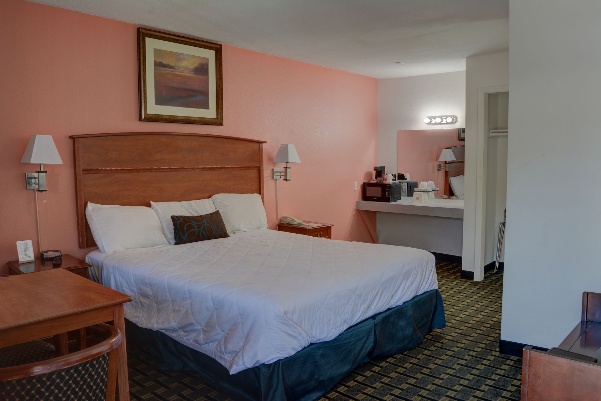 A motel room with a bed, a desk, and a vanity area against a peach-colored wall.