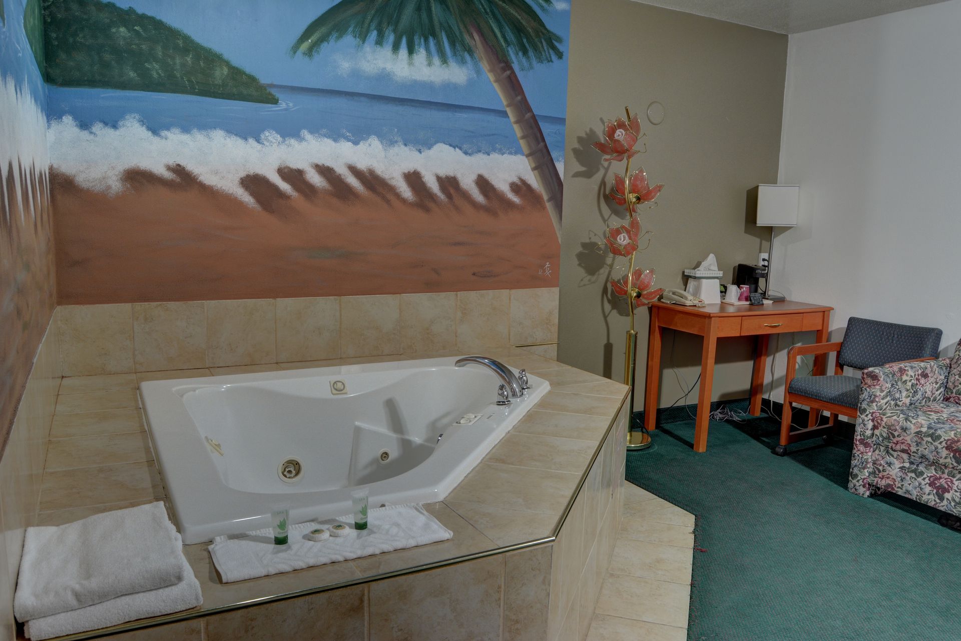 A hotel room with a jacuzzi tub, painted beach scene wall, small desk, and armchair on a teal carpet.