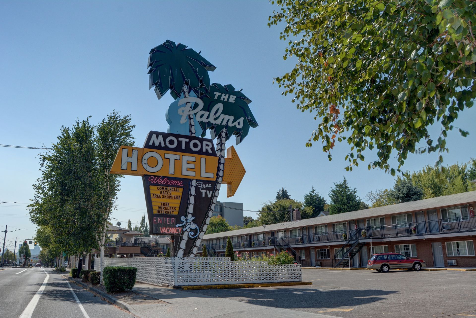 The Palms Motor Hotel sign with a large arrow, palm tree design, and a row of motel rooms on a sunny day.