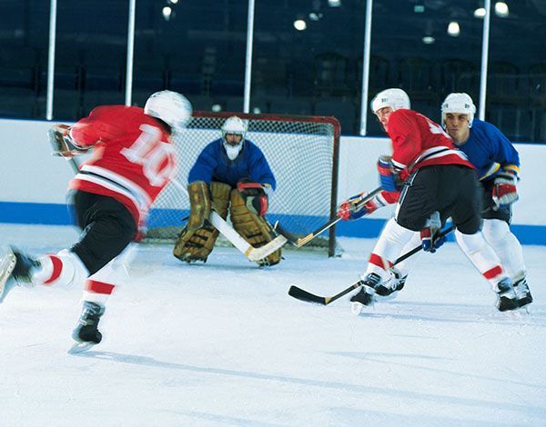 Hockey players in action, one shooting towards the goal. Red and blue jerseys clash on the ice rink, goalie in net.