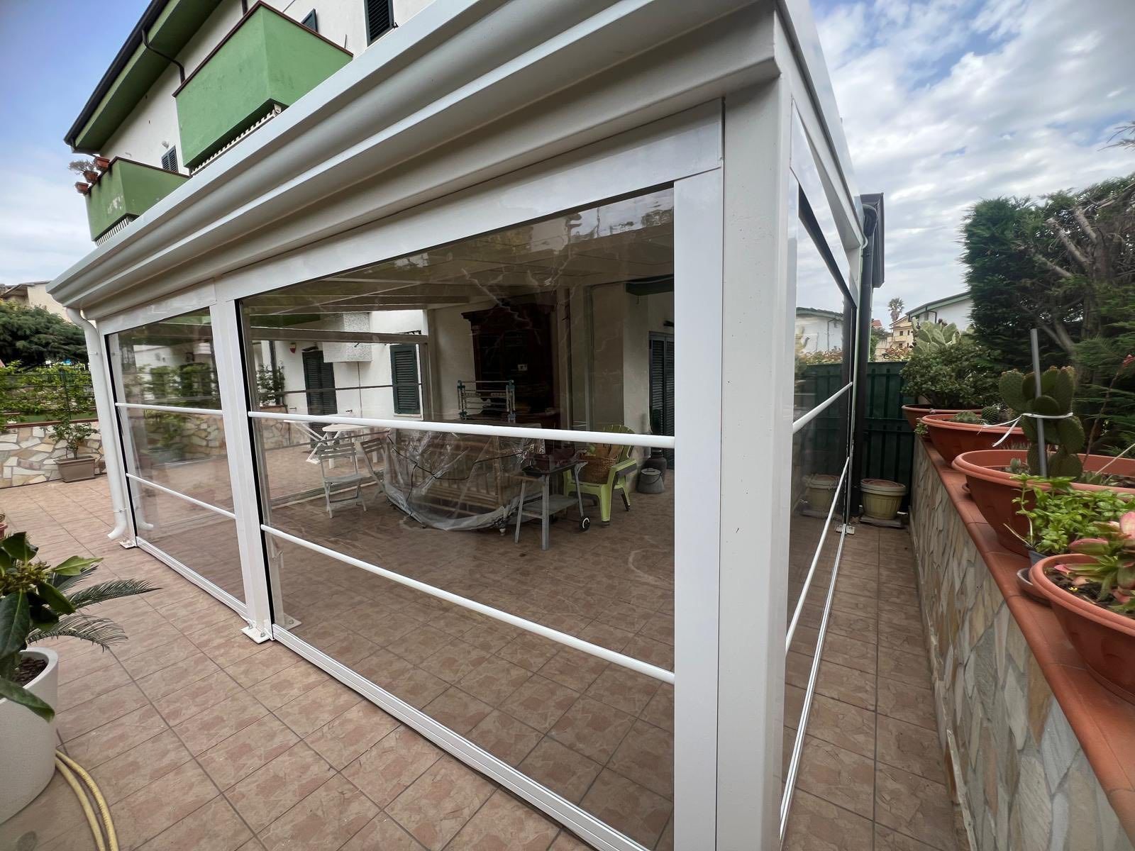 Un pergolato bianco con pareti in vetro trasparente si trova su un patio accanto a una casa.