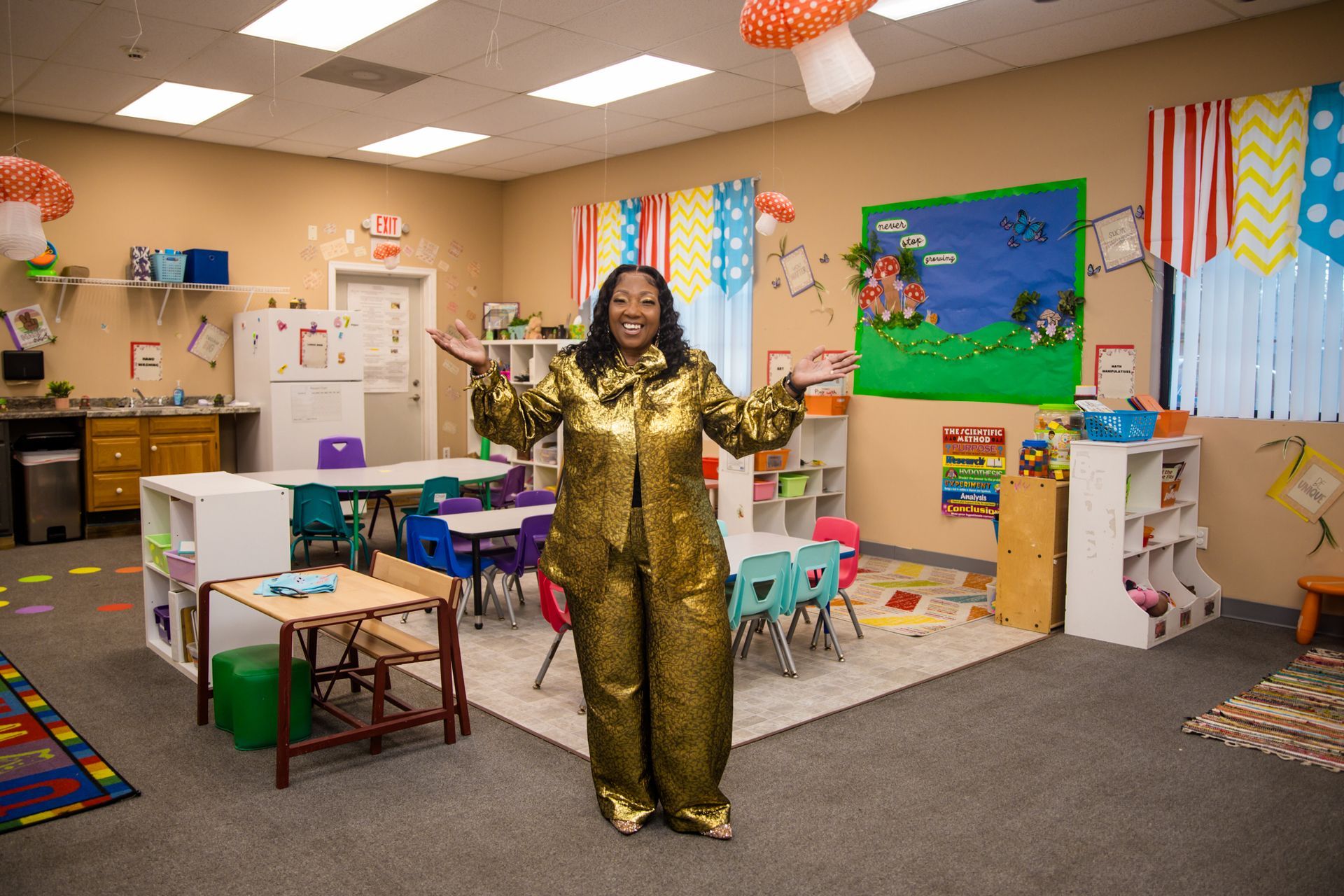 Woman in gold jumpsuit in a colorful preschool classroom with open arms, smiling.