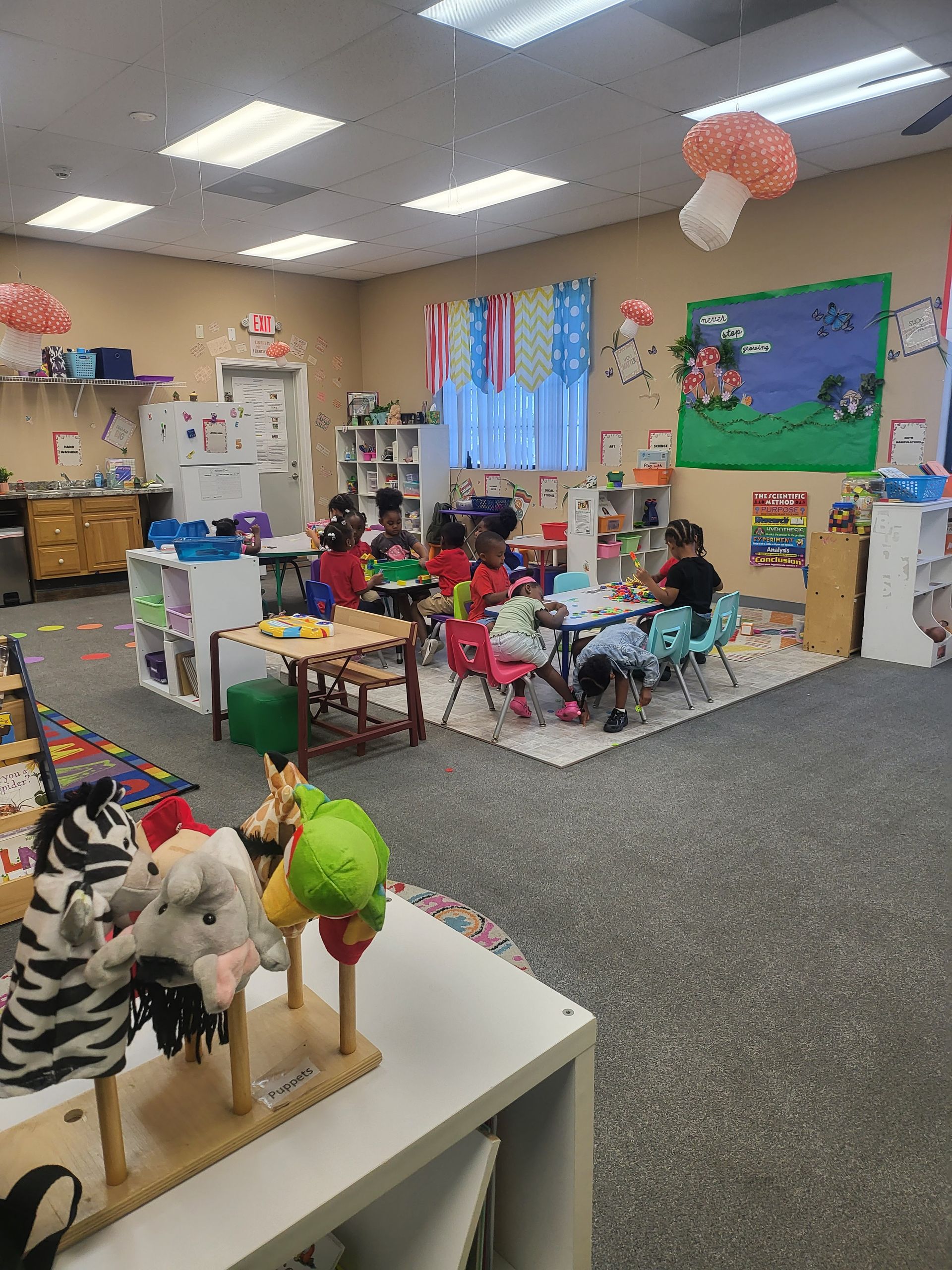 Children in a colorful classroom; several seated at tables, engaging in activities. Decorated room with puppets.