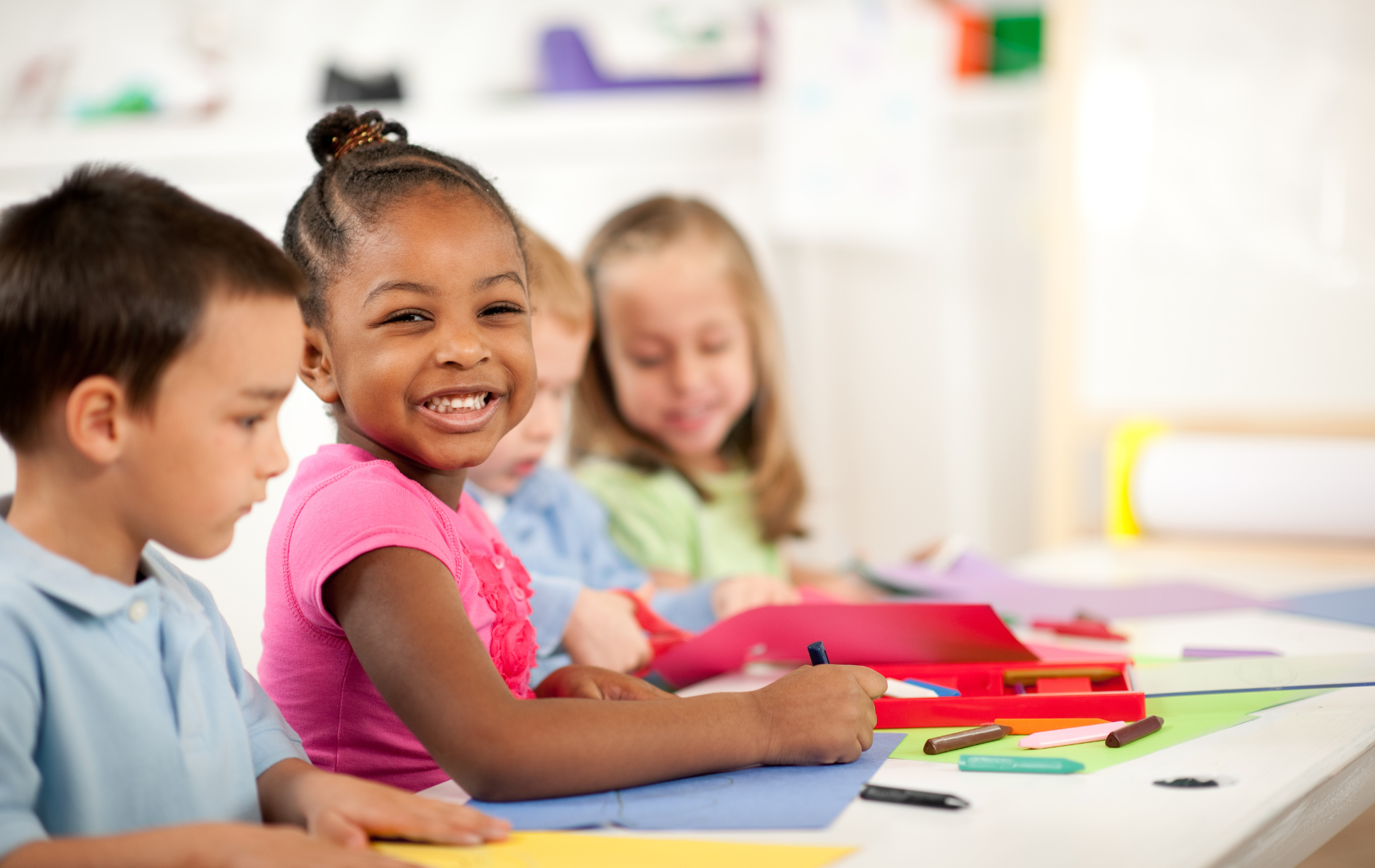 Children drawing with crayons at a table, smiling. Girl in pink shirt looks at camera.