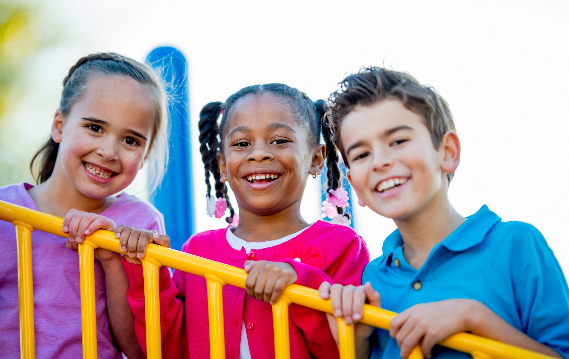 Three diverse children smiling at the camera, holding a yellow railing on a playground.